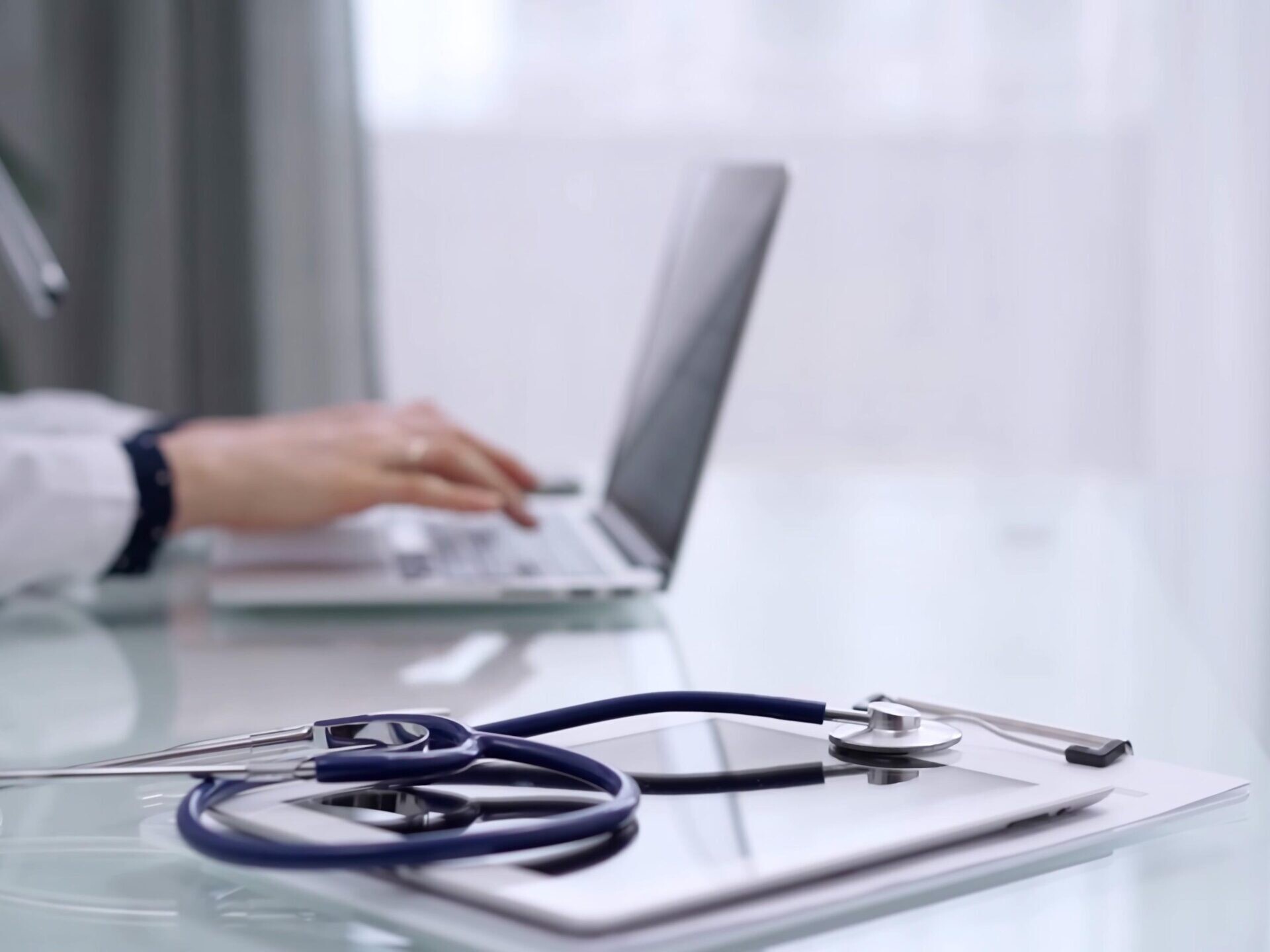 Hender som skriver på laptop i bakgrunnen. Stetoskop på notatblokk i forgrunnen