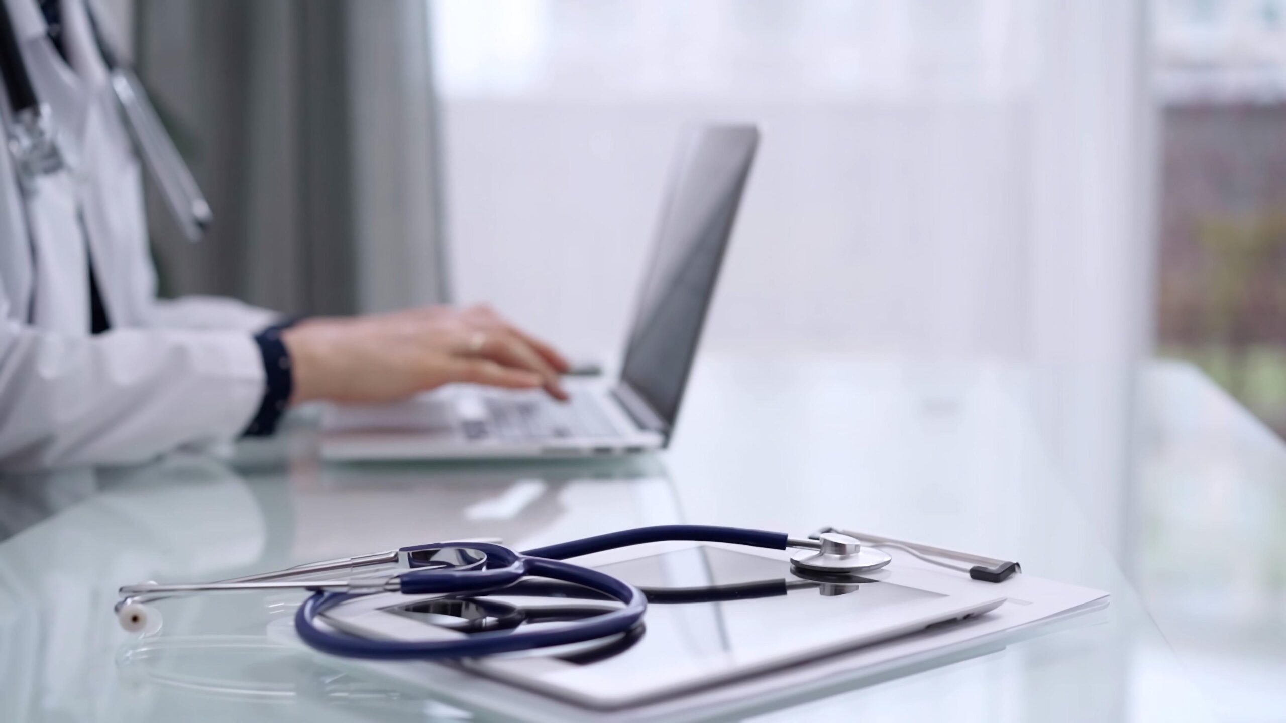 Hender som skriver på laptop i bakgrunnen. Stetoskop på notatblokk i forgrunnen