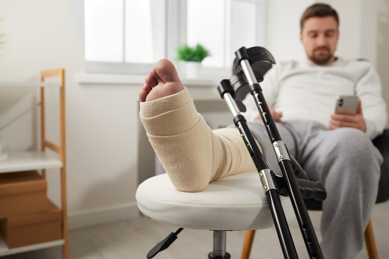 Fokus på gipset fot og krykker, mens vi ser uklart i bakgrunnen mannen som har den skadete foten